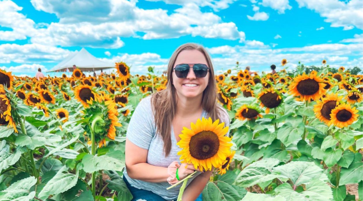A young woman with long blond hair wearing sunglasses and a gray tee shirt smiles as she holds a big, yellow sunflower at Barton Orchards in Poughquag.